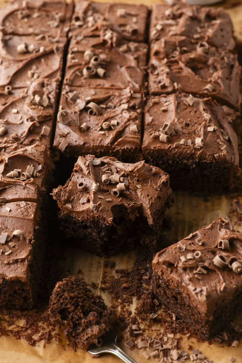 Several squares of chocolate cake with thick chocolate frosting and chocolate shavings are arranged on parchment paper, with one piece slightly removed and a forkful taken from it.