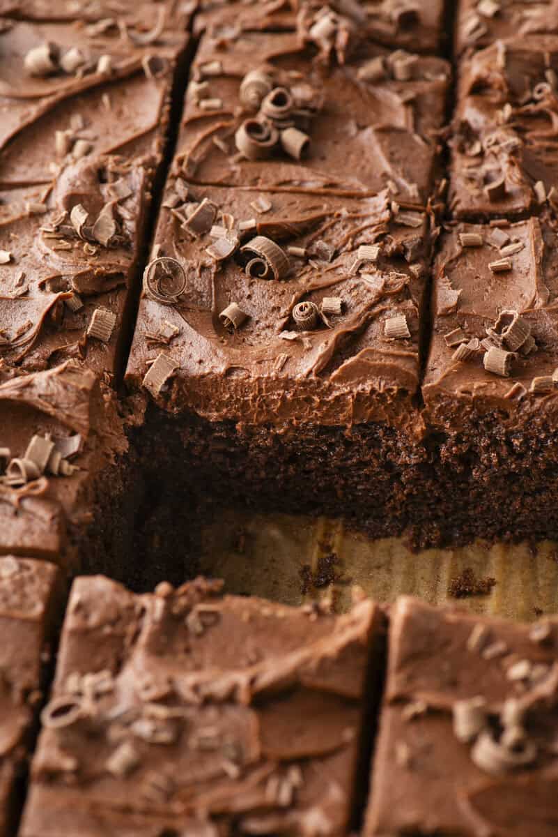 A close-up of sliced chocolate cake with chocolate frosting and chocolate shavings on top, with one piece missing from the tray.