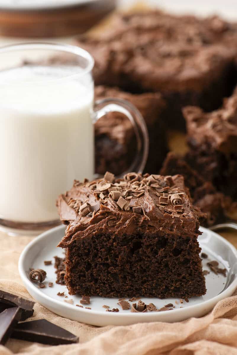 A slice of chocolate cake with chocolate frosting and shavings on a white plate, next to a glass of milk. More cake is visible in the background.