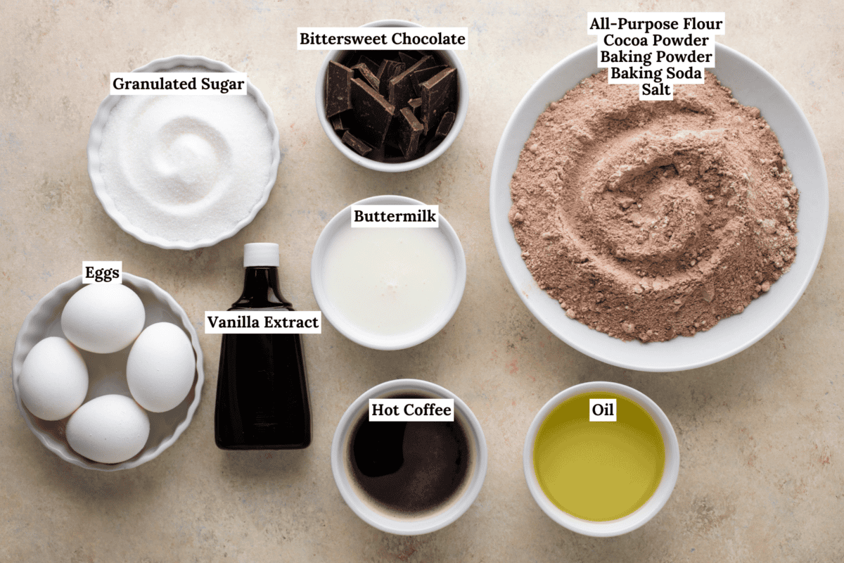 Overhead view of labeled baking ingredients in bowls: granulated sugar, bittersweet chocolate, eggs, vanilla extract, buttermilk, hot coffee, oil, and a flour mixture with cocoa powder, baking powder, baking soda, and salt.