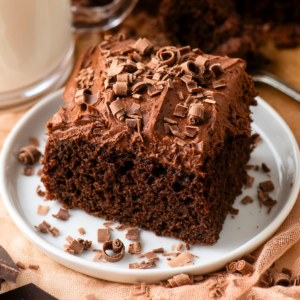 A square piece of chocolate cake with chocolate frosting and chocolate shavings on top, served on a white plate with more chocolate shavings scattered around.
