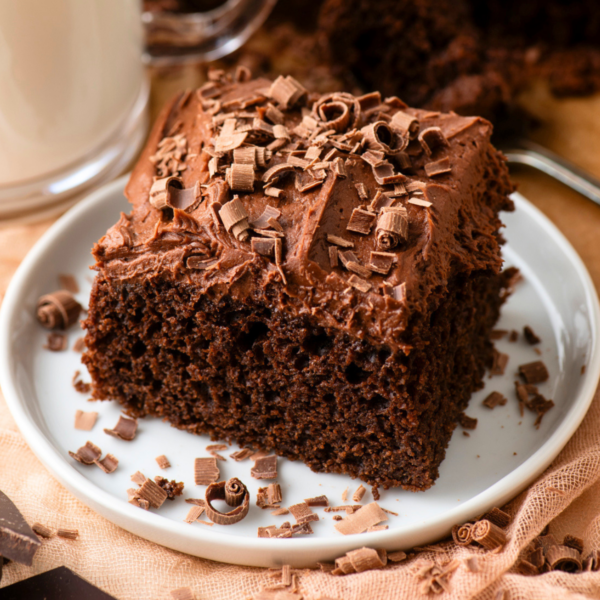 A square piece of chocolate cake with chocolate frosting and chocolate shavings on top, served on a white plate with more chocolate shavings scattered around.