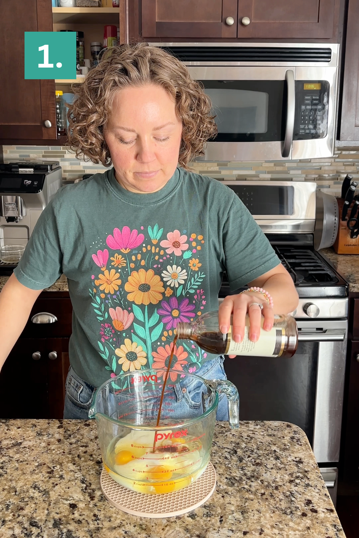 A person with short curly hair and a floral shirt pours vanilla extract into a Pyrex measuring cup with eggs in a kitchen. A teal square with 1. is in the top left corner.