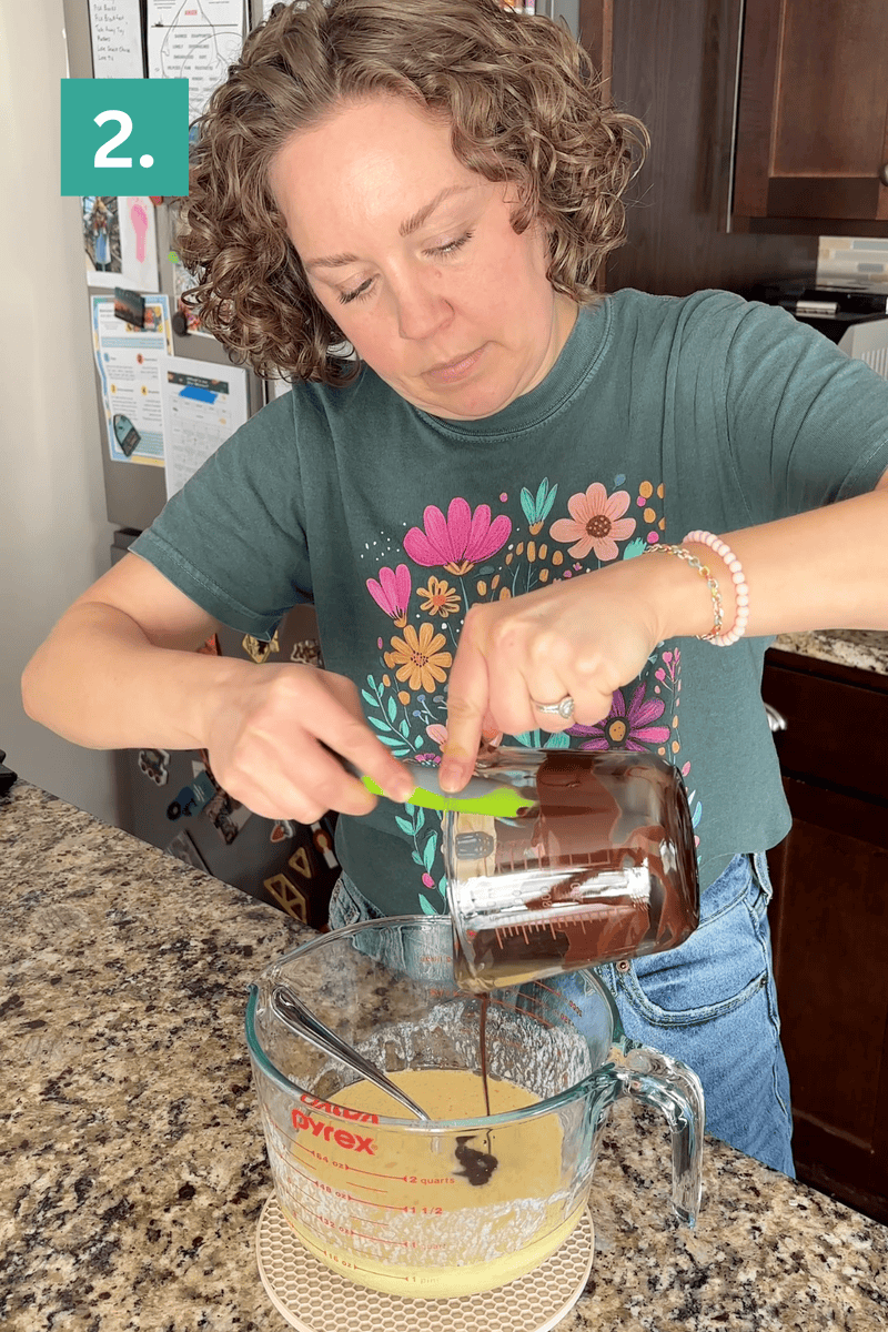 A woman with curly hair pours melted chocolate from a glass measuring cup into another mixing cup on a kitchen counter. She wears a green floral shirt and jeans. A green box with the number 2. is in the top left corner.