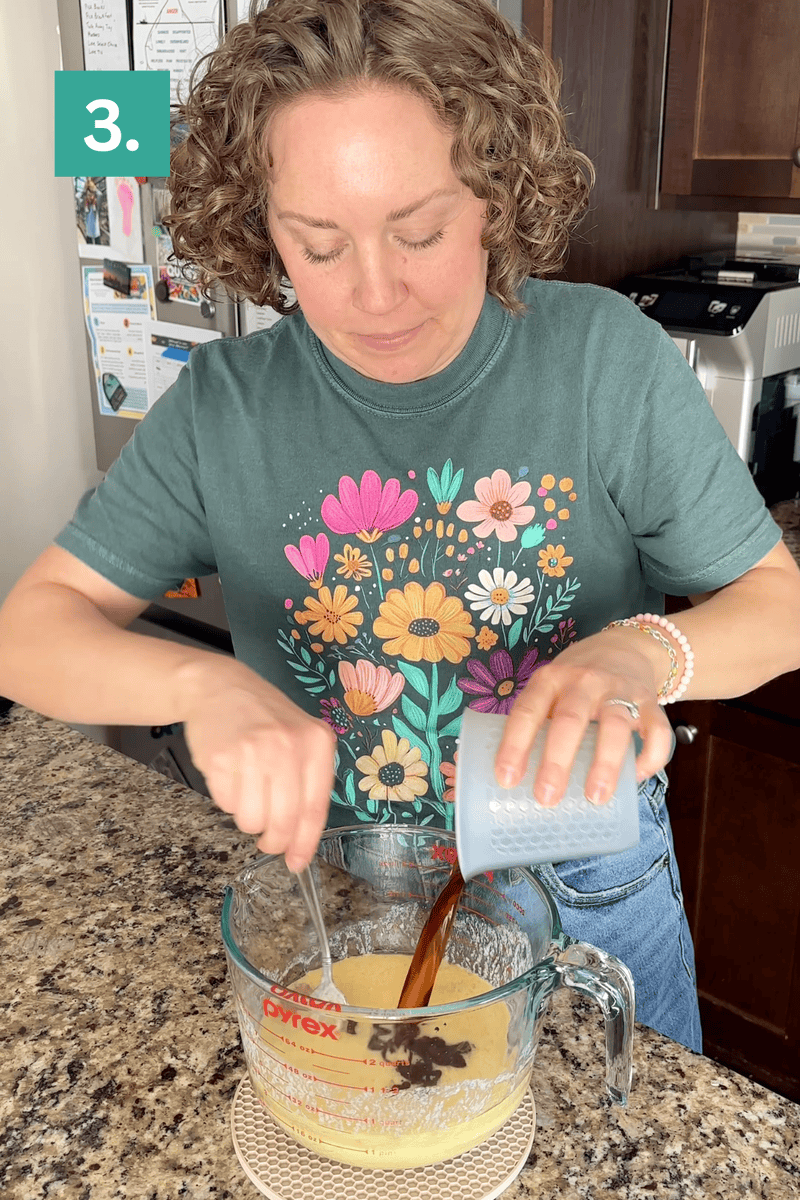 A person with curly hair, wearing a floral t-shirt, stands at a kitchen counter, pouring a dark liquid from a small cup into a large Pyrex measuring cup while stirring the mixture. A green square with the number 3 is in the top left corner.