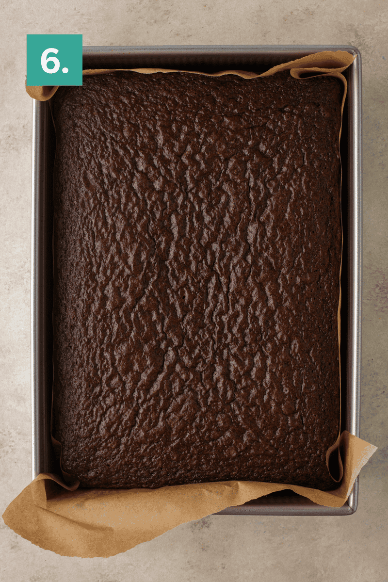 A freshly baked chocolate cake in a square pan lined with parchment paper, viewed from above. A green box with the number 6 appears in the top left corner.