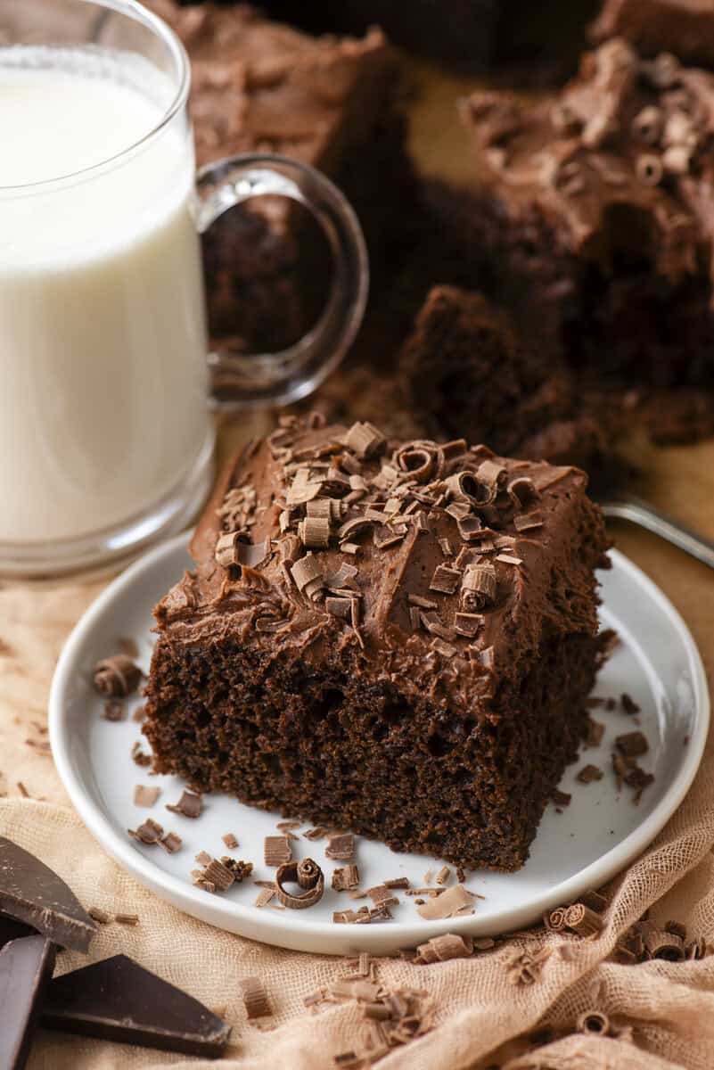 A slice of chocolate cake with chocolate frosting and chocolate shavings sits on a white plate next to a glass of milk. Chocolate pieces and more shavings are scattered around on the table.