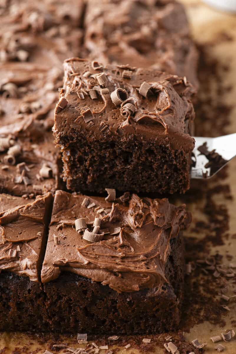 A close-up of a square piece of chocolate cake with thick chocolate frosting and chocolate shavings being lifted from a tray of similar pieces on parchment paper.