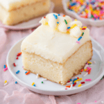 A square slice of vanilla cake with white frosting, decorated with piped frosting and colorful sprinkles, sits on a white plate with more sprinkles scattered around.