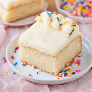 A square slice of vanilla cake with white frosting, decorated with piped frosting and colorful sprinkles, sits on a white plate with more sprinkles scattered around.