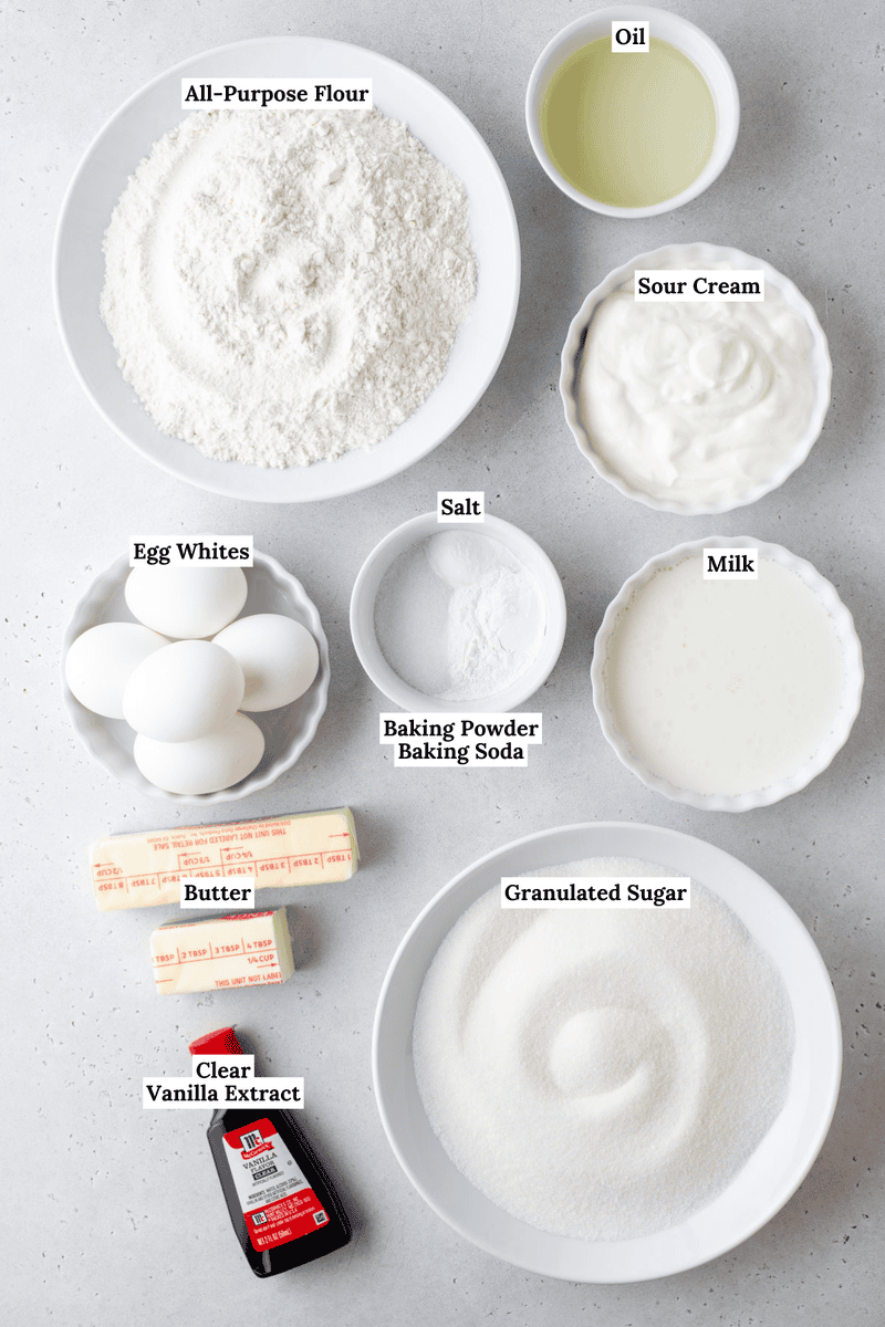 Top-down view of labeled cake ingredients in bowls and plates, including all-purpose flour, oil, sour cream, egg whites, salt, baking powder, baking soda, milk, butter, granulated sugar, and clear vanilla extract.