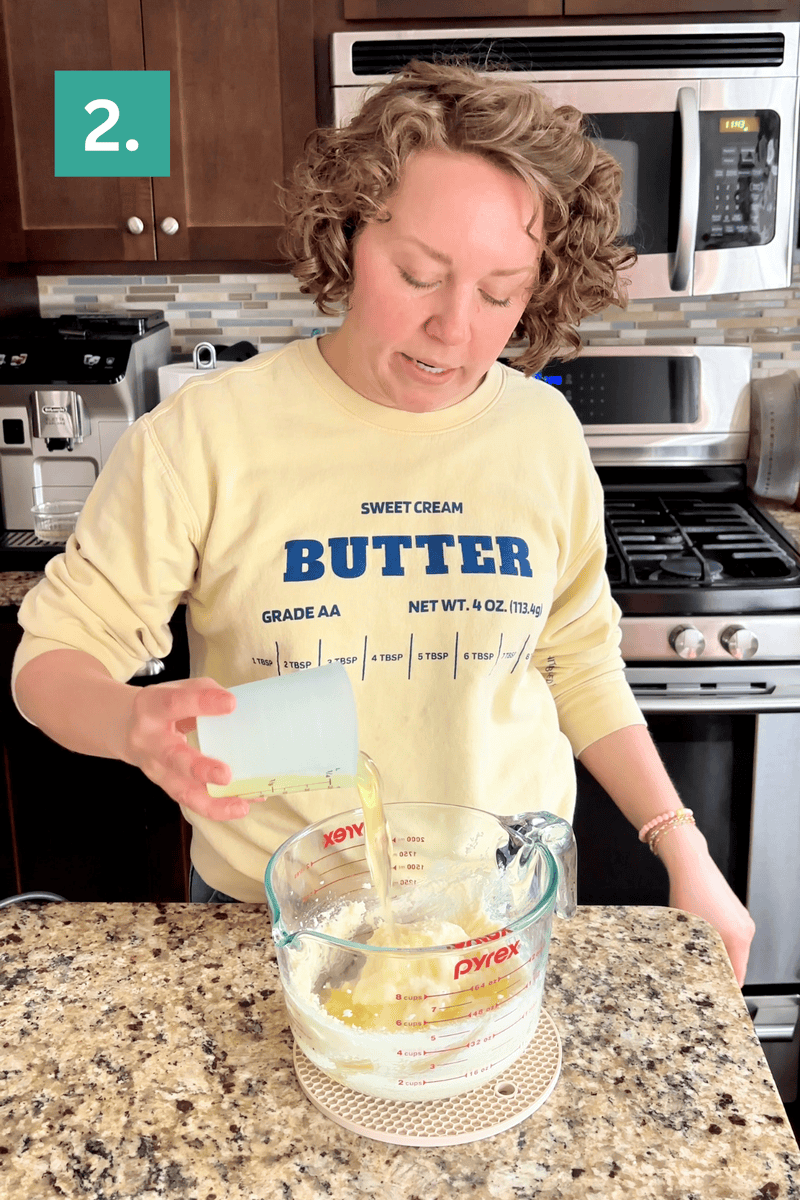 A person pours oil into a large glass measuring cup filled with ingredients on a kitchen counter. They wear a yellow BUTTER sweatshirt and stand in front of a stove and microwave. A teal box with 2. is in the top left corner.