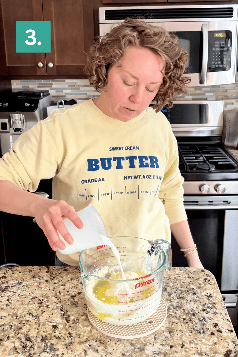 A person in a yellow Butter sweatshirt pours milk from a small cup into a glass measuring pitcher with melted butter on a kitchen counter. A stove and microwave are visible in the background. A green box labeled 3. is in the top left corner.
