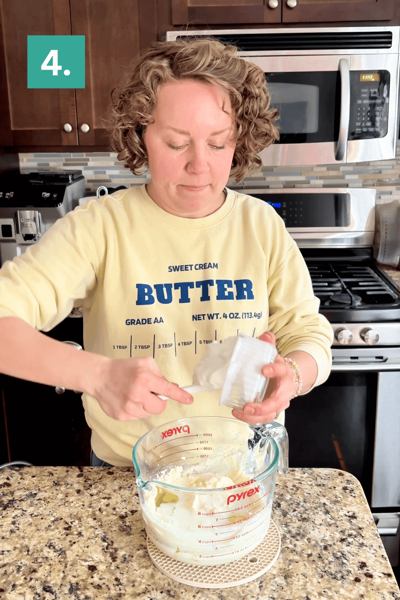 A person adds an ingredient from a small container into a glass measuring cup with a mixture inside, standing in a kitchen. A green square with the number 4 is in the upper left corner.