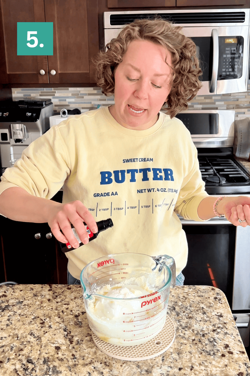 A person wearing a yellow BUTTER sweatshirt stands in a kitchen, pouring vanilla extract from a bottle into a glass Pyrex measuring cup on a granite countertop. A green step 5 label appears in the corner.