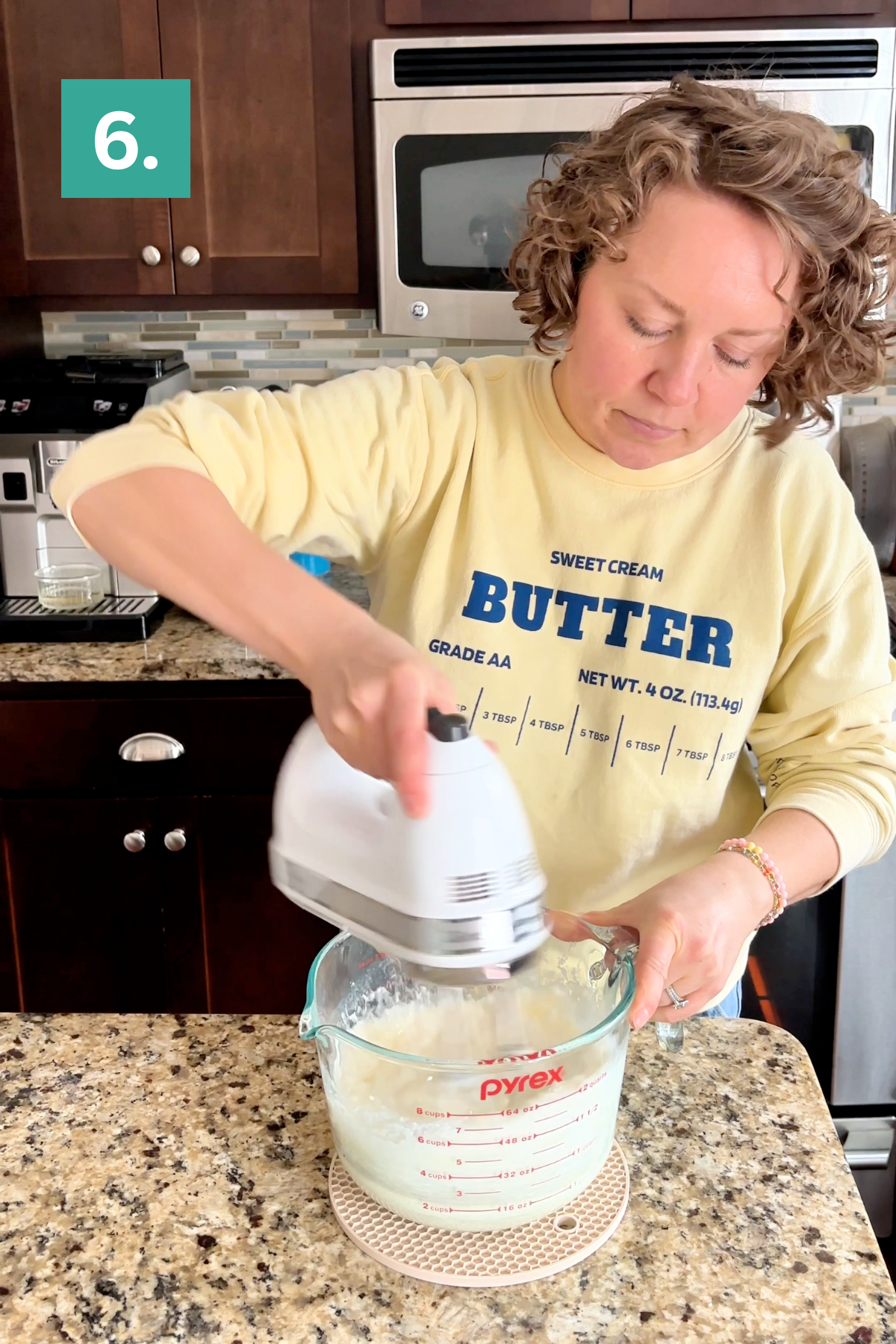 A woman wearing a yellow Butter sweatshirt uses a handheld mixer to blend ingredients in a Pyrex measuring cup on a kitchen counter. A teal box with the number 6 is in the corner.