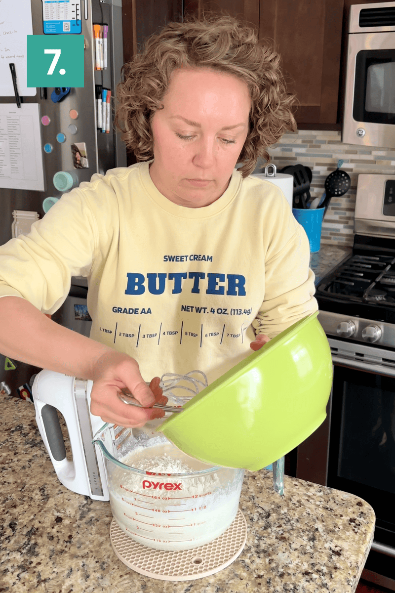 A person in a yellow Butter sweatshirt pours ingredients from a green bowl into a glass measuring cup on a kitchen counter, with a hand mixer and kitchen appliances visible in the background.