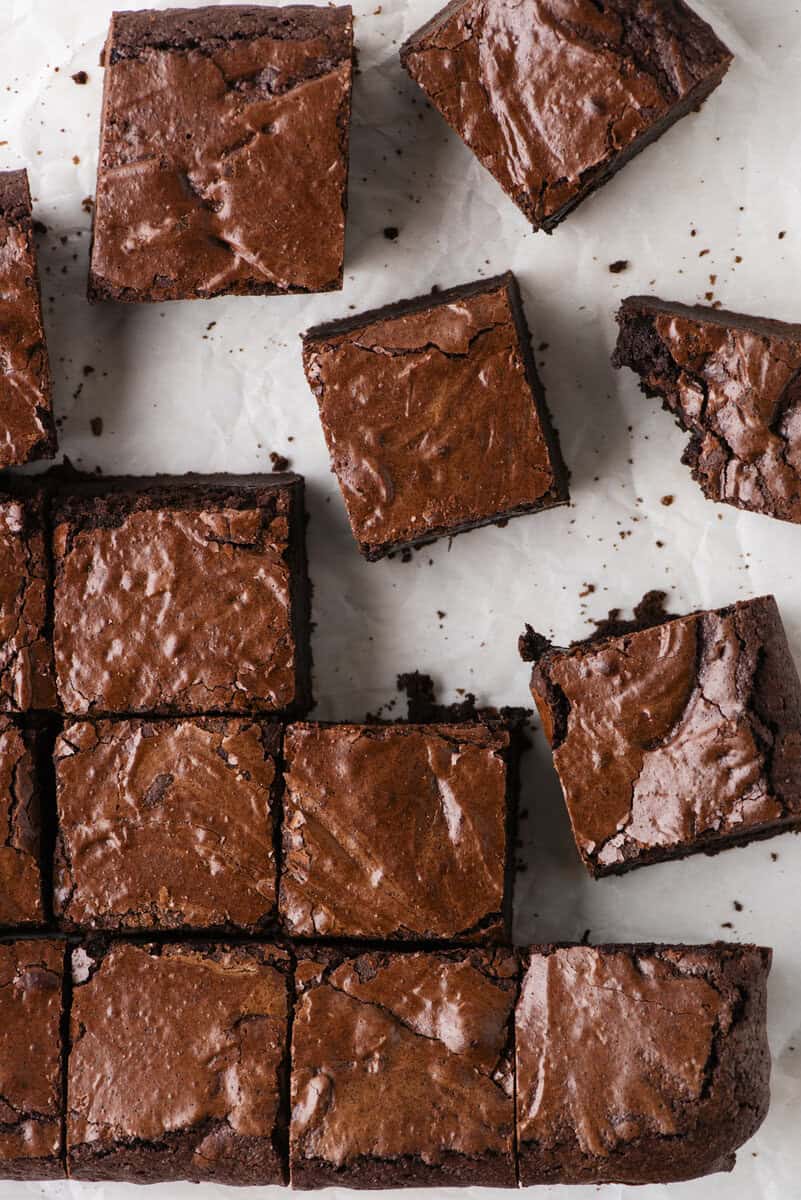 Squares of chocolate brownies with a shiny, crackled top are arranged on white parchment paper, some pieces separated and a couple with bites taken out. The brownies appear rich, fudgy, and freshly baked.