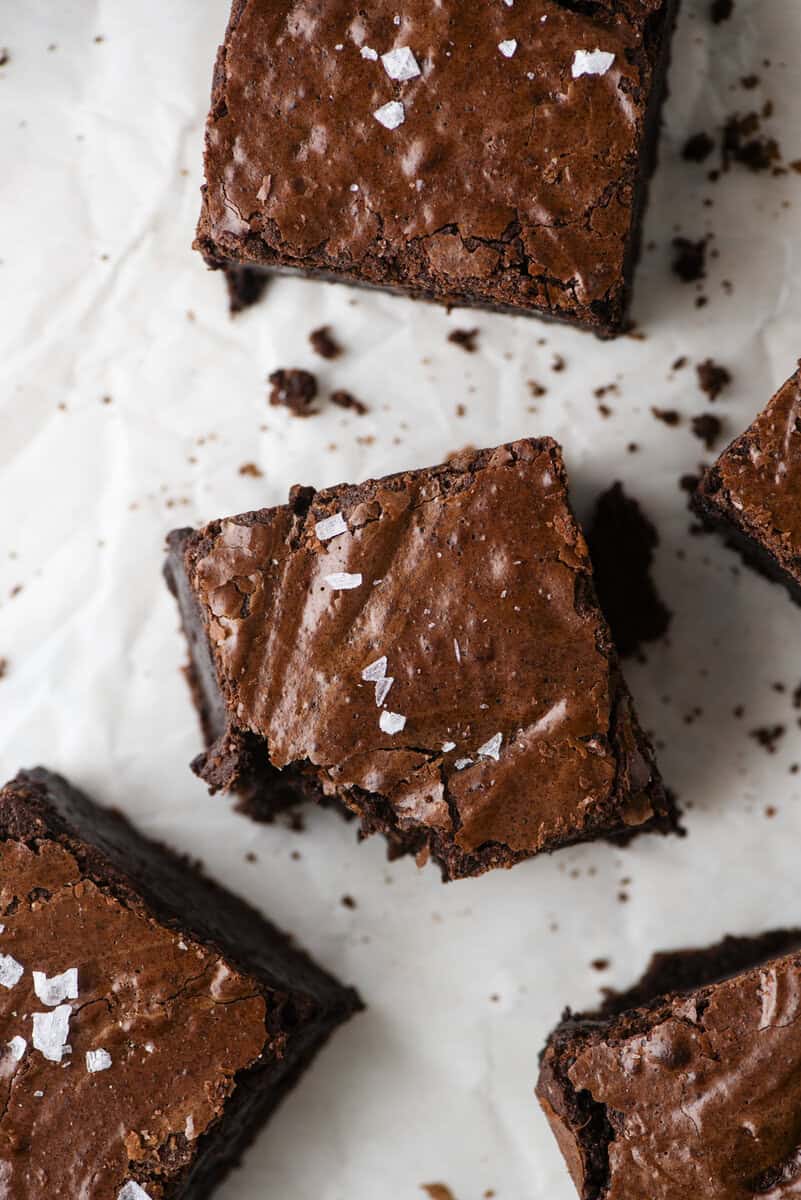 Three fudgy chocolate brownies topped with flaky sea salt are arranged on white parchment paper. One brownie has a bite taken out of it, showing its moist and rich interior.
