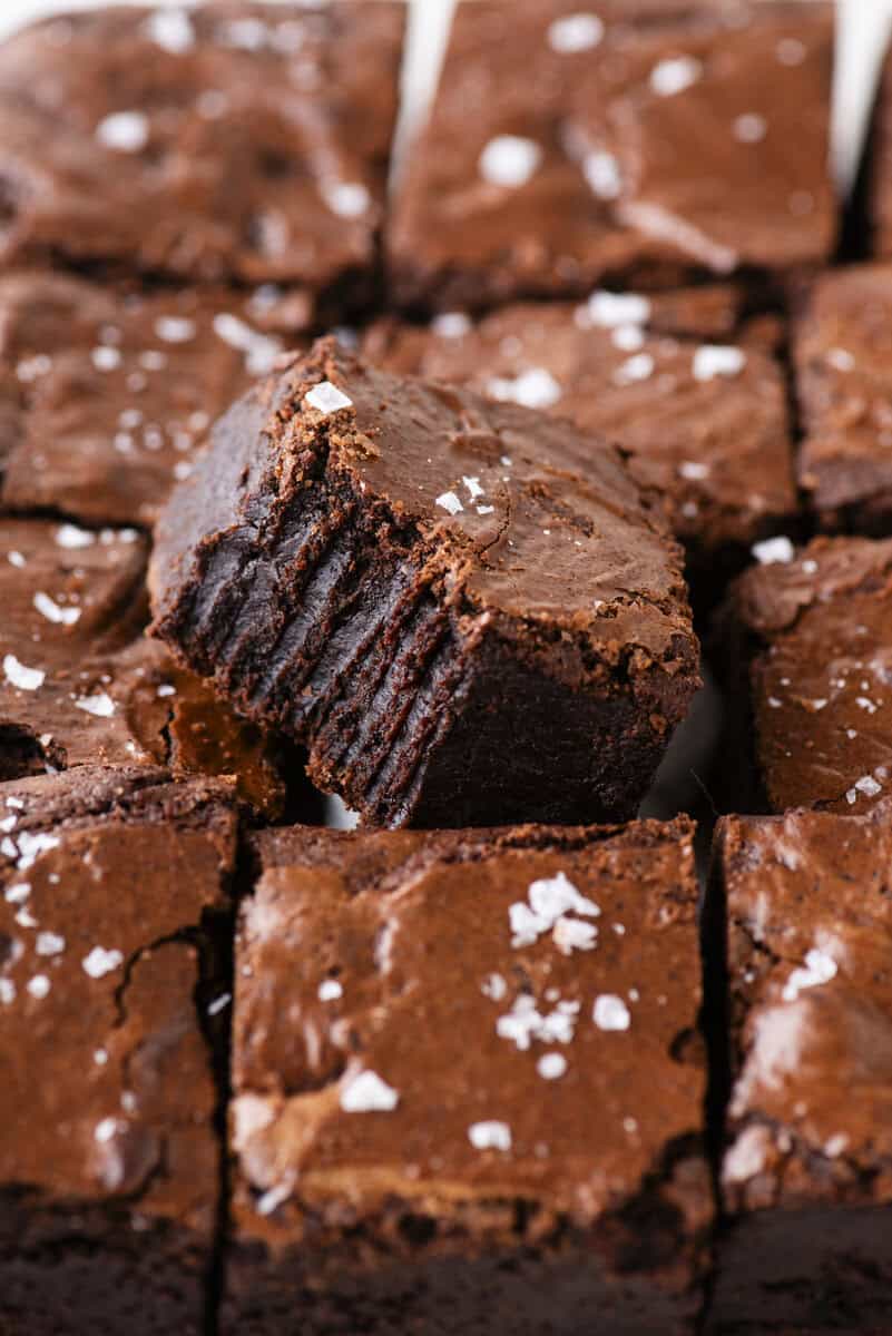 A batch of fudgy chocolate brownies cut into squares, with one brownie on top showing bite marks. The brownies are topped with a sprinkle of flaky sea salt.