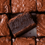 Close-up of thick, fudgy brownies with a shiny, cracked top. One brownie is turned on its side to show its dense, moist texture, surrounded by other neatly cut brownies.