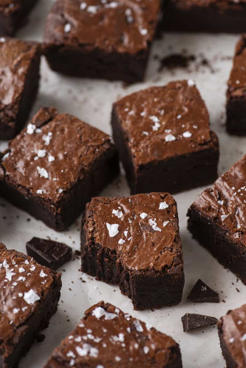 Squares of rich, fudgy brownies topped with flaky sea salt are arranged on parchment paper, with one brownie showing a bite taken out and small chocolate pieces scattered around.