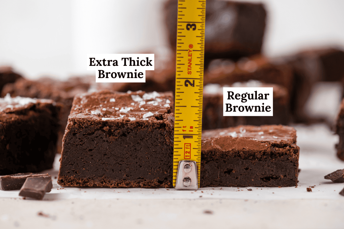 A side-by-side comparison of an extra thick brownie and a regular brownie with a tape measure showing the extra thick brownie is about 2 inches tall. Labels identify each brownie, and there are chocolate pieces nearby.