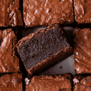 Close-up of thick, fudgy brownies with a shiny, cracked top. One brownie is turned on its side to show its dense, moist texture, surrounded by other neatly cut brownies.