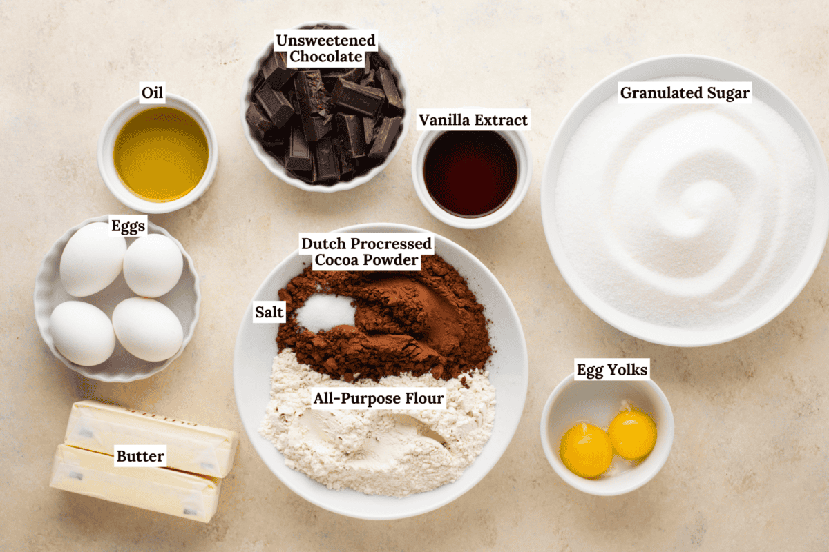 A top-down view of bowls and plates containing labeled baking ingredients: eggs, butter, oil, unsweetened chocolate, vanilla extract, granulated sugar, all-purpose flour, Dutch processed cocoa powder, salt, and egg yolks.