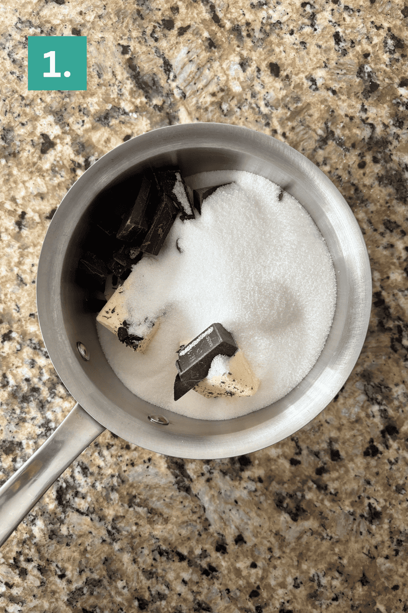 A small saucepan on a granite countertop contains sugar, chunks of dark chocolate, and cubes of butter. A green box with the number 1 is in the top left corner.
