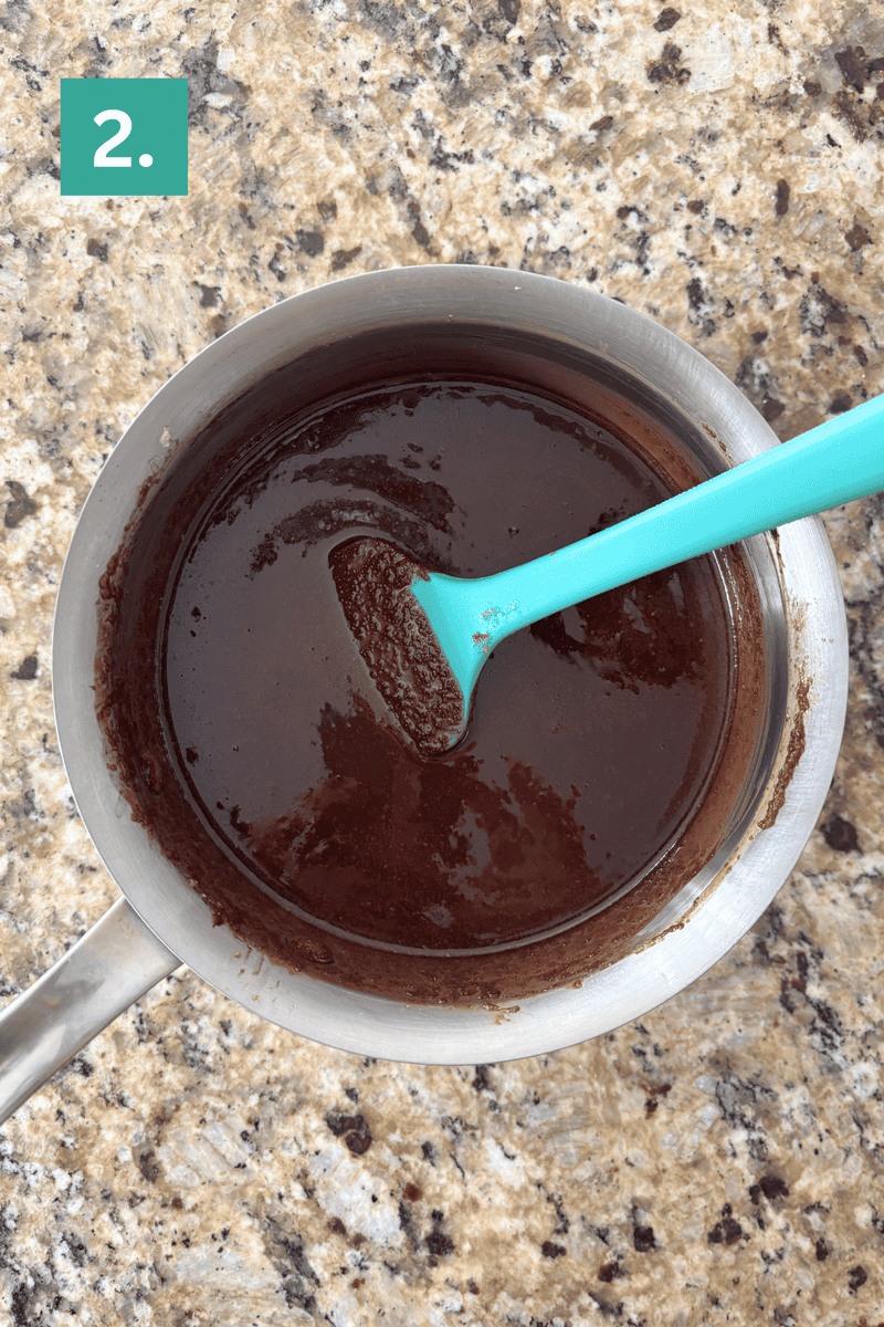 A saucepan with smooth, melted chocolate mixture being stirred by a teal spatula, placed on a speckled granite countertop. A green box with the number 2. is in the top left corner.