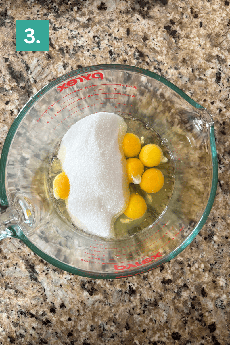 A glass mixing cup on a speckled countertop contains six eggs and a mound of sugar. A green box labeled 3. appears in the top left corner, indicating step three of a recipe.