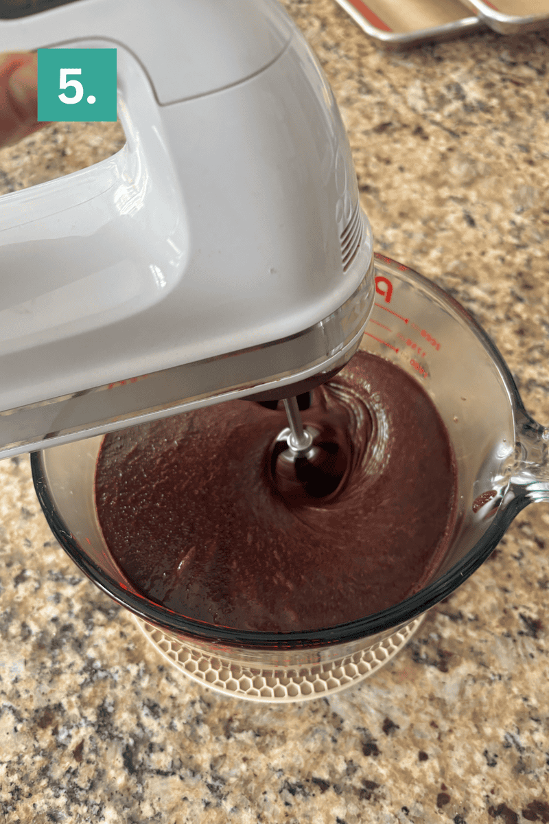 A hand mixer blends chocolate batter in a glass measuring cup on a granite countertop. A green square with the number 5 is in the top left corner.