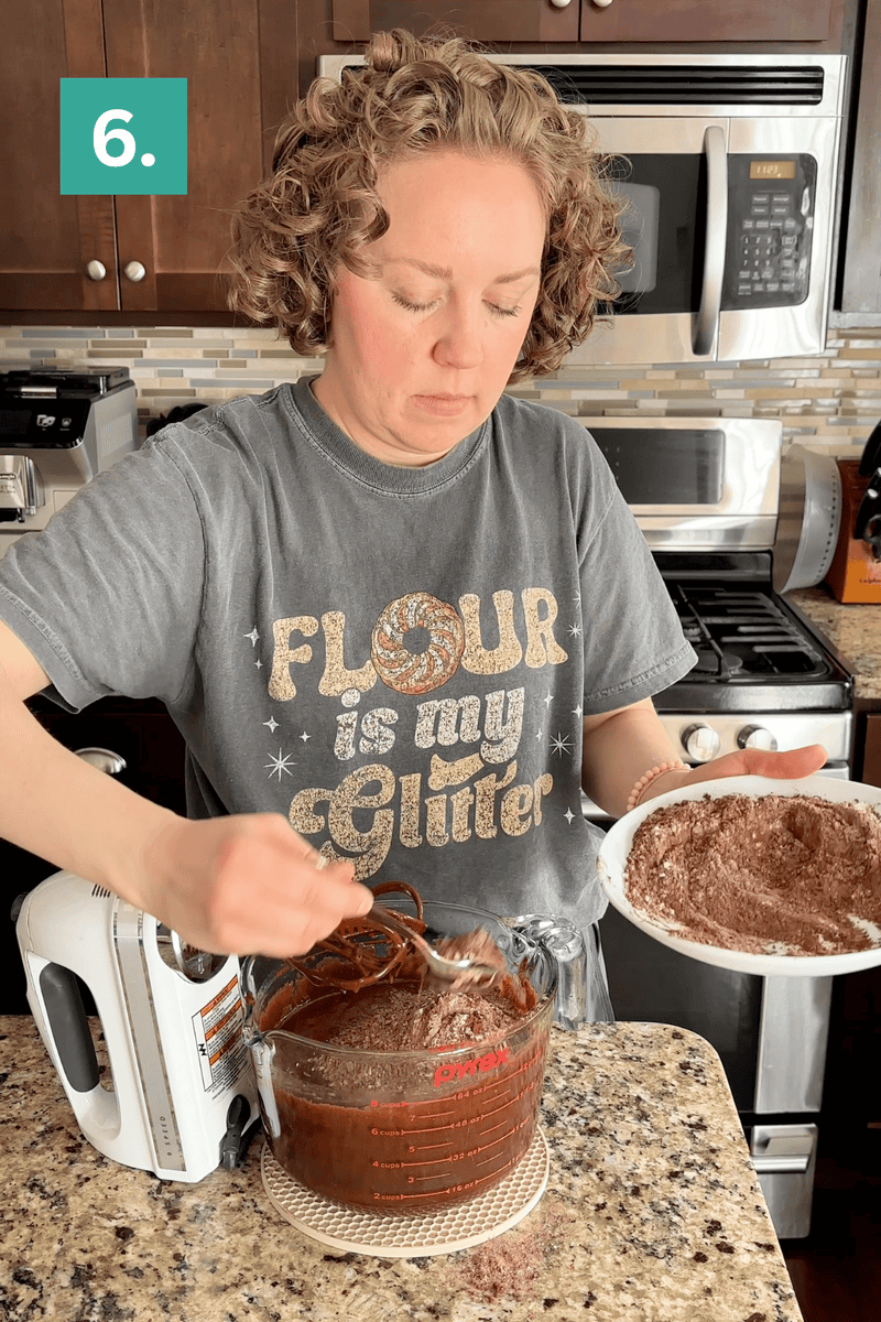 A woman in a kitchen mixes chocolate cake batter in a glass bowl with a hand mixer while holding a plate of dry ingredients. She wears a shirt that says Flour is my glitter. A green label with the number 6 appears in the corner.