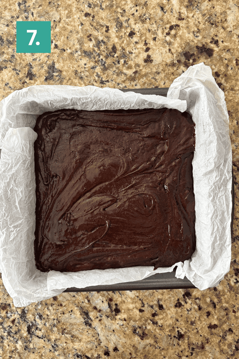 A baking pan lined with parchment paper is filled with smooth, unbaked chocolate batter. The pan sits on a granite countertop. In the upper left corner, a green box labeled “7.” is visible.