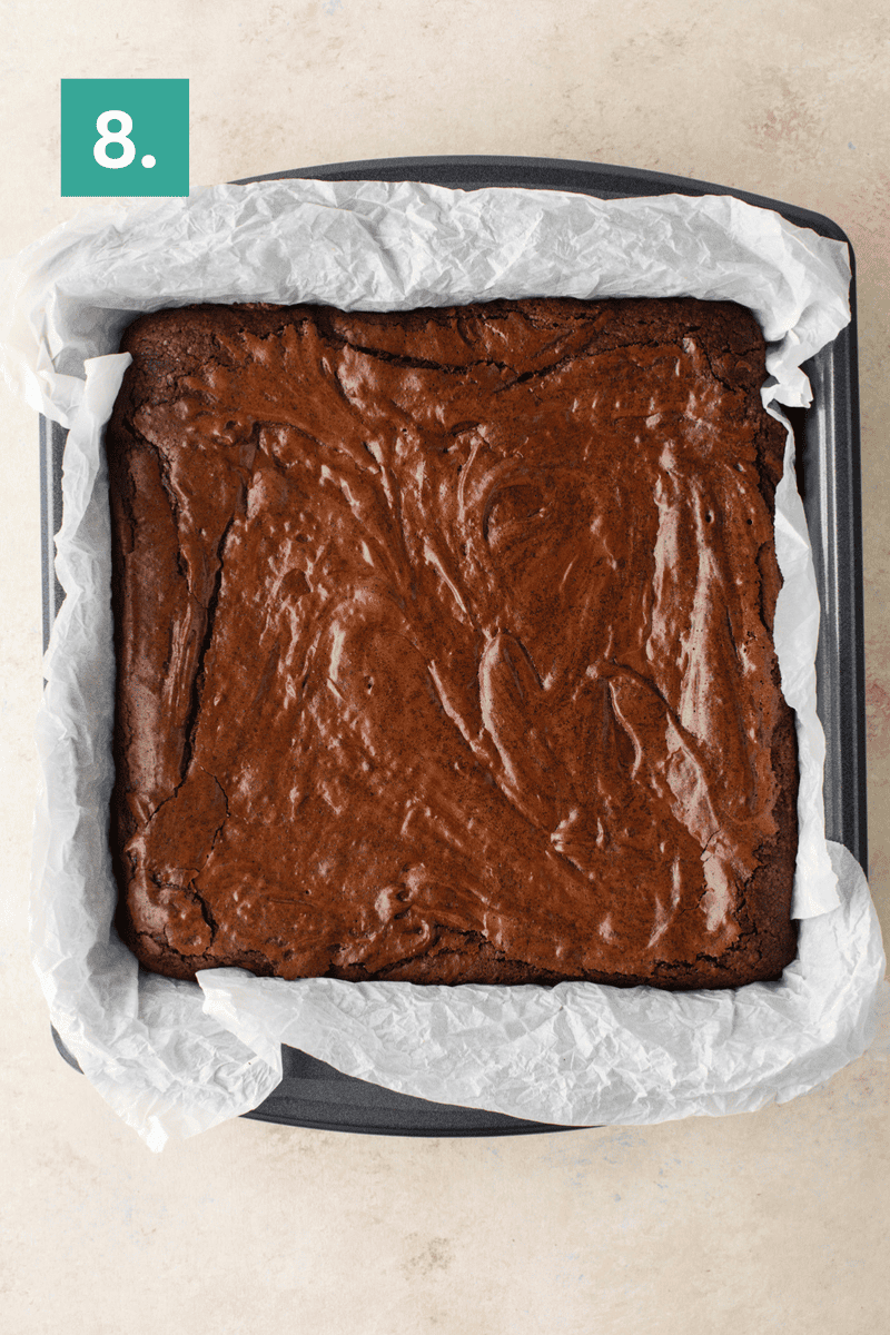 A square baking pan lined with parchment paper holds freshly baked brownies with a glossy, crackled top. A green box in the top left corner displays the number 8.