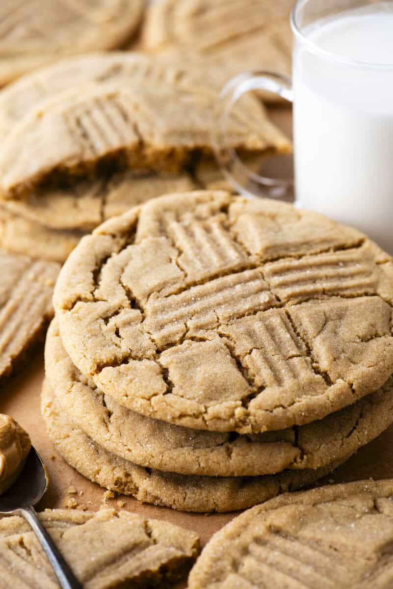 A close-up of several peanut butter cookies stacked and scattered on parchment paper, with a glass of milk in the background and a spoonful of peanut butter nearby. The cookies have a classic crisscross fork pattern.