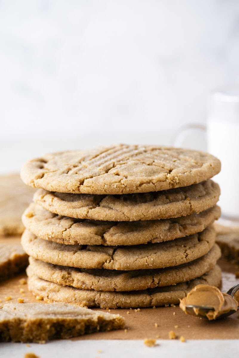 A stack of seven golden brown peanut butter cookies sits on parchment paper, with crumbs and a spoonful of peanut butter nearby. A glass of milk is blurred in the background.