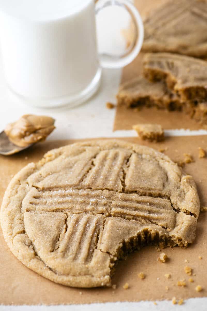 A peanut butter cookie with a bite taken out sits on parchment paper, next to a spoonful of peanut butter, cookie pieces, and a glass of milk in the background.