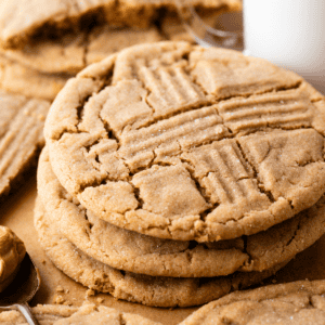 A stack of peanut butter cookies with a crisscross fork pattern sits on parchment paper next to a spoonful of peanut butter and a glass of milk.