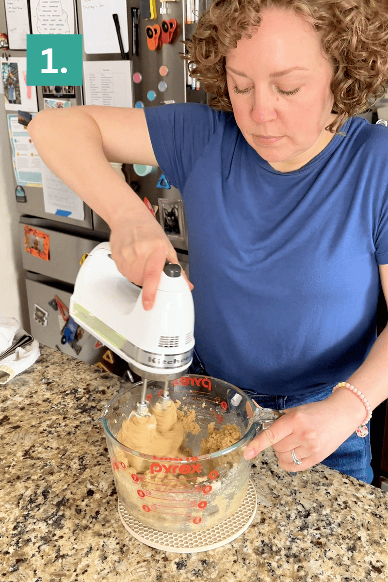 A woman in a blue shirt uses a hand mixer to blend ingredients in a large glass measuring cup on a granite kitchen counter; a refrigerator with magnets is in the background.