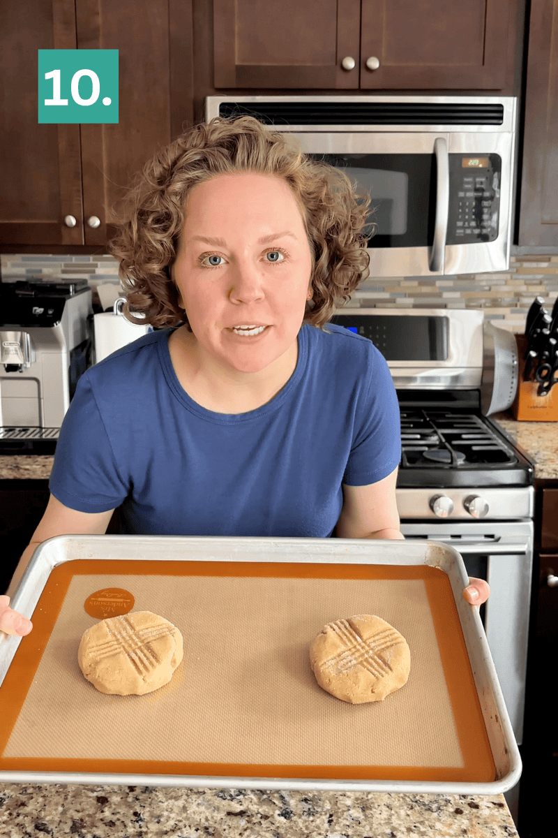 A woman with curly hair, wearing a blue shirt, holds a baking tray with two unbaked peanut butter cookies in a kitchen. A green box with 10. is in the top left corner of the image.