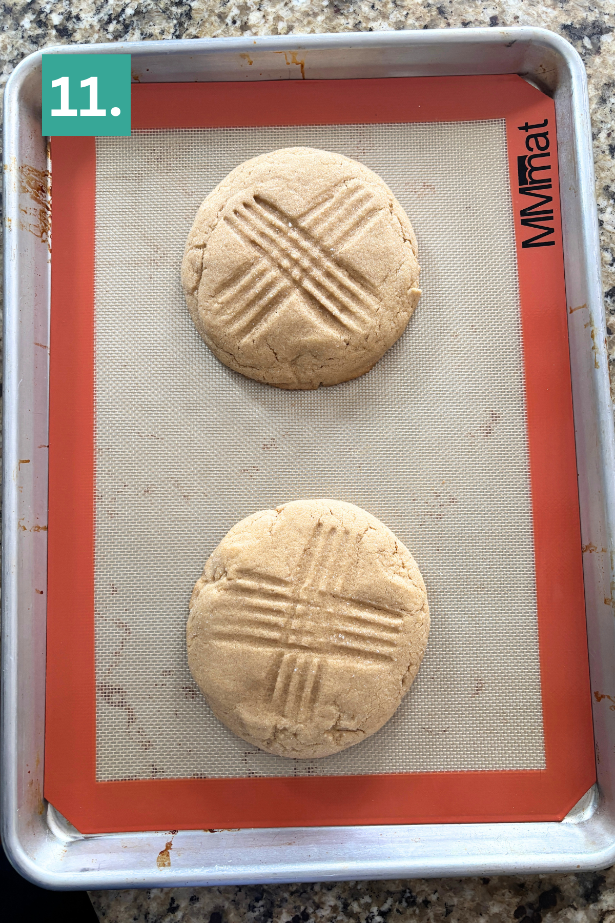 Two large, round cookies with crisscross fork marks on top are on a silicone baking mat-lined tray. The tray is on a granite countertop, and a green label with 11. is in the top left corner.
