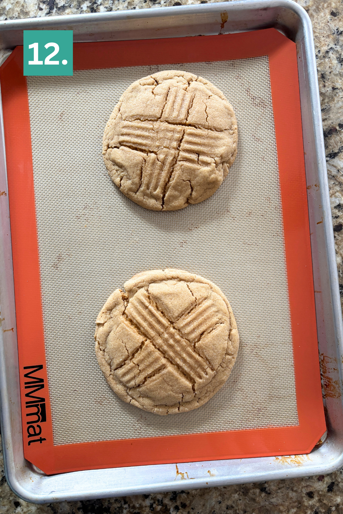 A baking tray with a silicone mat holds two round cookies, each with a crisscross pattern on top. A green square with the number 12. is in the top left corner of the image.
