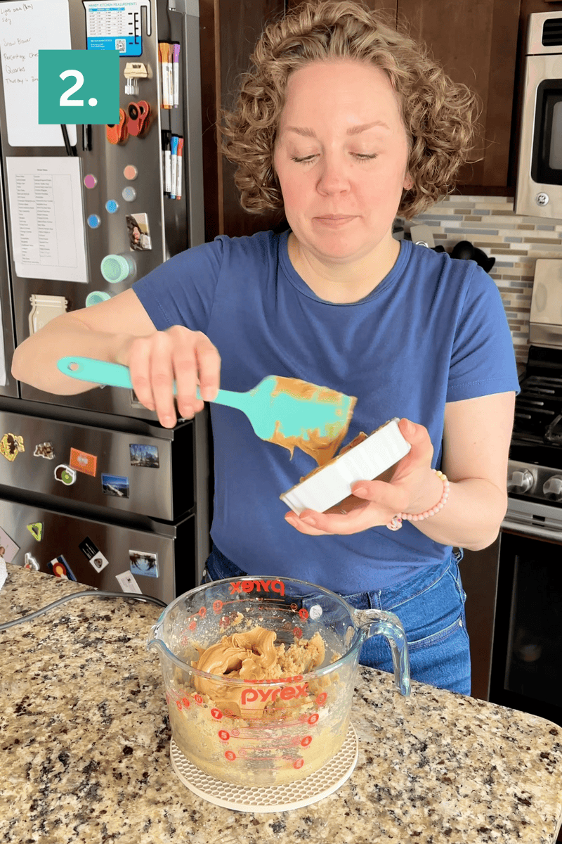 A woman in a blue shirt uses a teal spatula to scoop peanut butter from a measuring cup into a white container in a kitchen, with a fridge and stove in the background. A 2. label is in the top left corner.