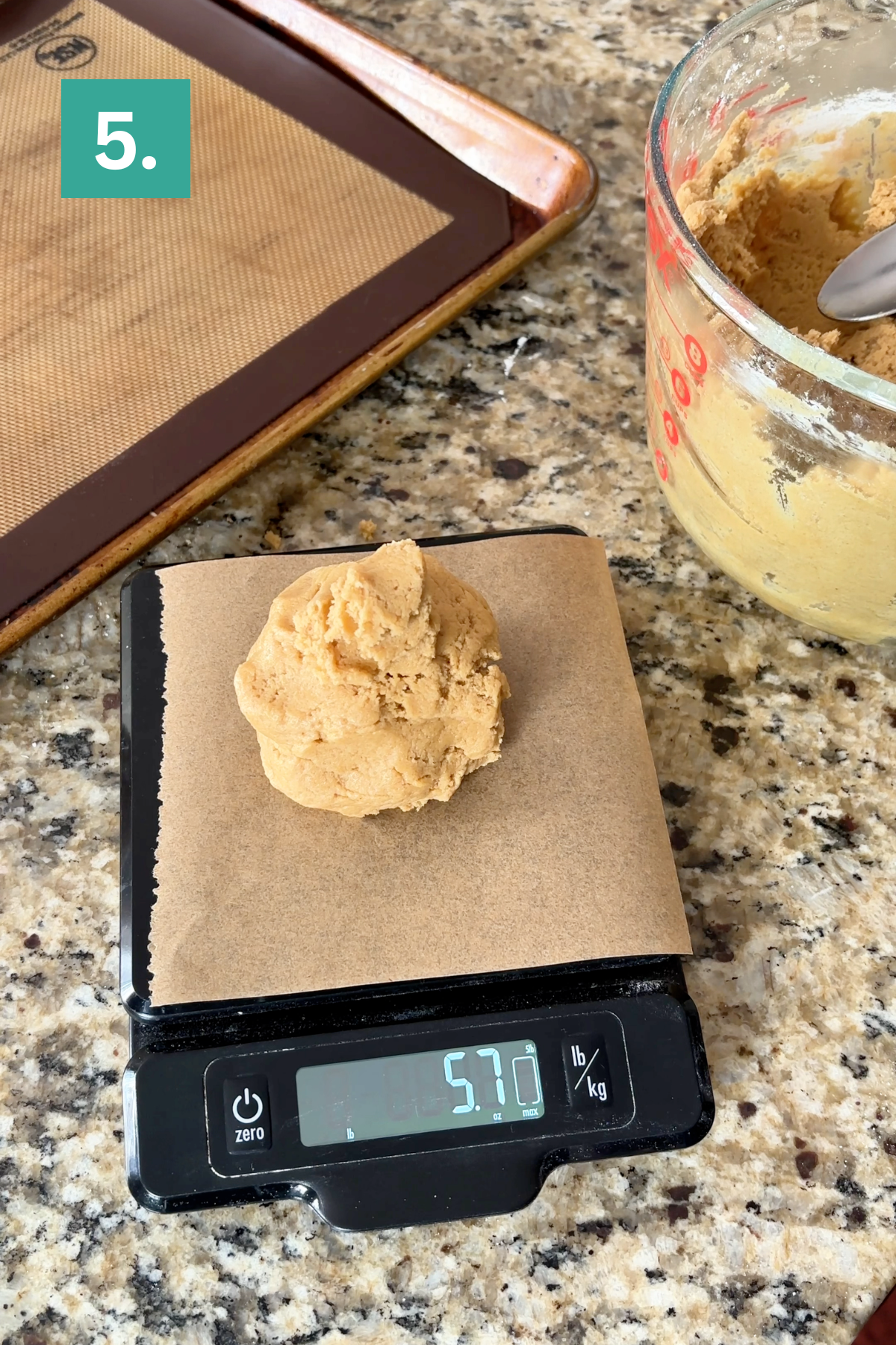 A scoop of cookie dough sits on parchment paper atop a digital scale reading 57.0 grams. Nearby are a lined baking sheet and a mixing bowl with dough on a granite countertop. A green label with 5. is at the top left.