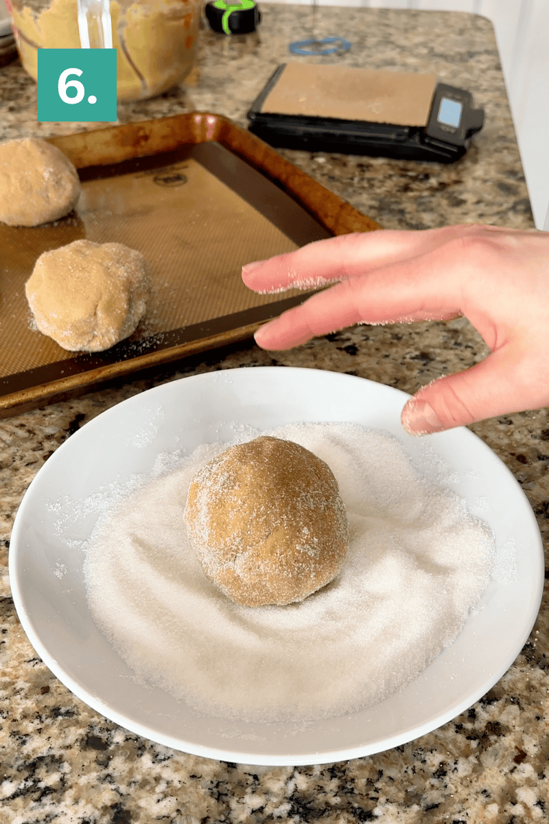 A hand rolls a ball of cookie dough in a plate of sugar. Another dough ball is on a baking tray lined with a silicone mat. A kitchen scale and more dough are visible in the background. A green box with “6.” is in the top left corner.