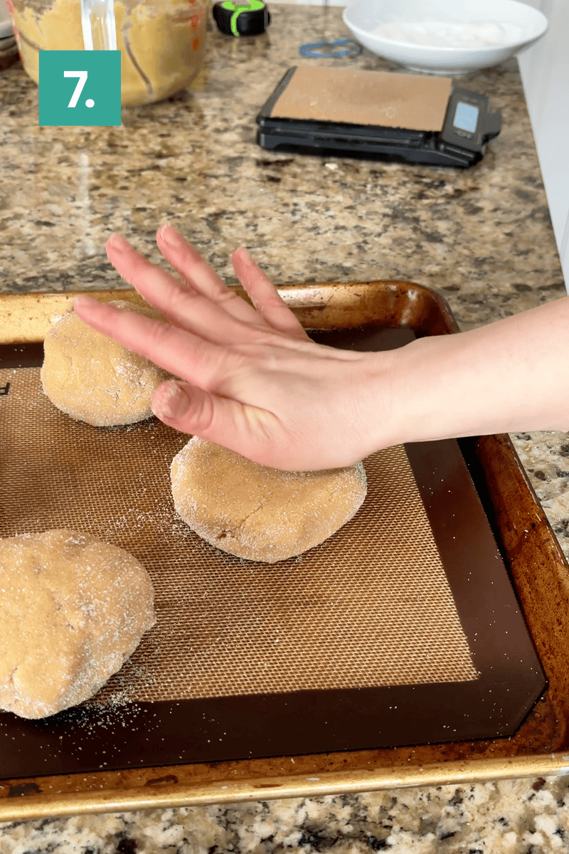 A hand presses down on a ball of dough, flattening it on a baking sheet lined with a silicone mat. Two other dough balls are nearby, and baking ingredients and tools are in the background. A green box in the top left reads “7.”.