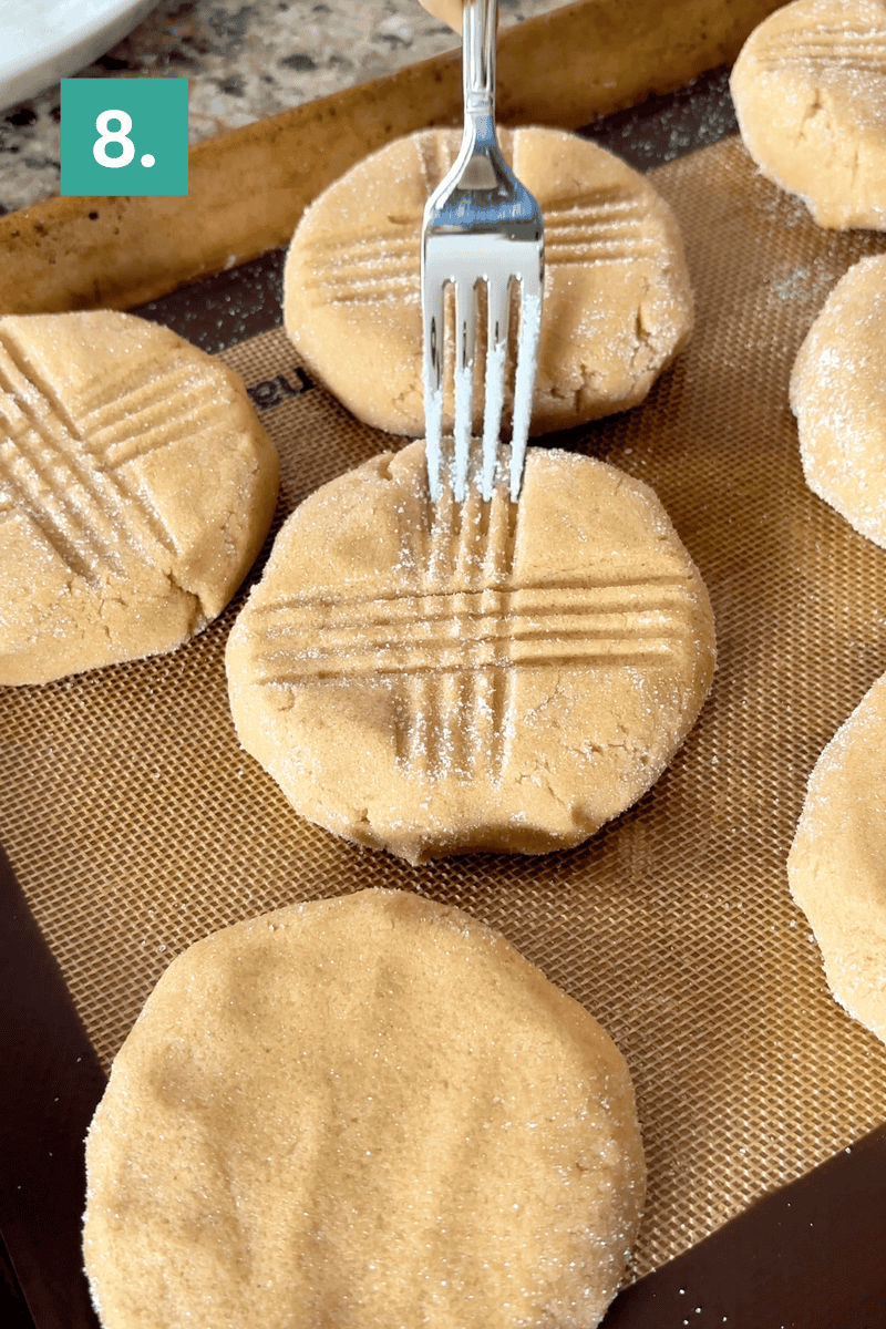 A fork presses a crisscross pattern into raw, round cookie dough on a baking sheet lined with a silicone mat. Other sugar-coated cookies are nearby. A teal box with the number 8 is in the top left corner.
