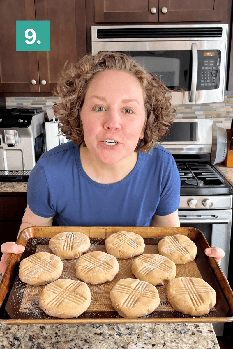 A person with curly hair and a blue shirt holds a baking tray with ten round, crisscross-topped cookies in a kitchen. A green box with the number 9 is in the upper left corner.